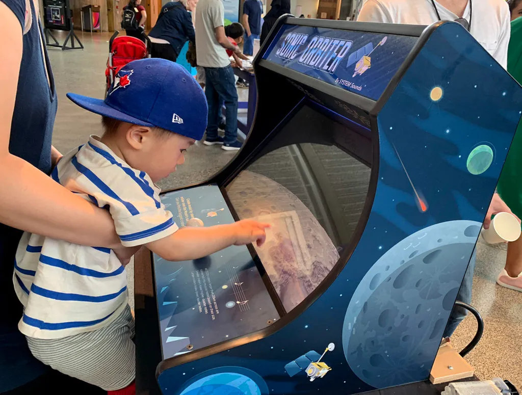 A very young child being held by his mother while he touches the screen of the Sonic Orbiter